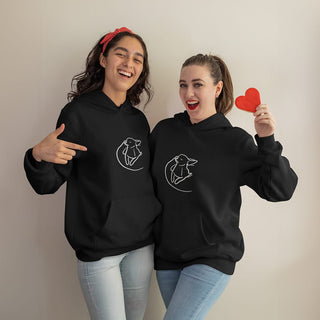 Two joyful friends wearing black hoodies with custom embroidered pet portraits, one holding a heart-shaped cutout.