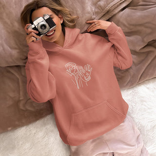 Person relaxing in a blush pink hoodie with custom hand-sign embroidery, playfully covering their face with a vintage camera.