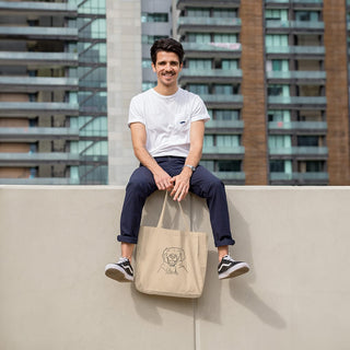 Cheerful person seated on a wall holding a Maison Memoire tote bag with a custom line-drawn portrait, cityscape in the background.