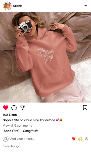 Sophia, a beaming bride-to-be, is snuggled in a cozy pink Maison Memoire hoodie with a custom engagement illustration, capturing her excitement.