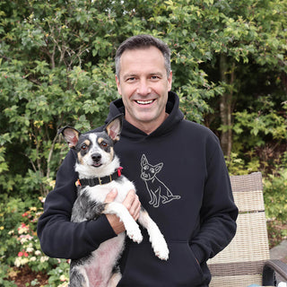 Smiling man wearing a black hoodie with a line drawing of a dog, holding a mixed breed dog with a black and white coat and pointy ears.