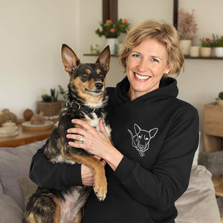 Smiling woman wearing a black hoodie with a line drawing of a dog, holding a mixed breed dog with pointy ears.