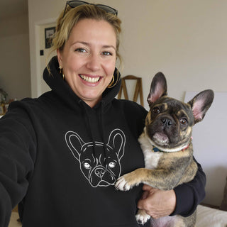 Smiling woman wearing a black hoodie with a line drawing of a French Bulldog, holding a real French Bulldog.