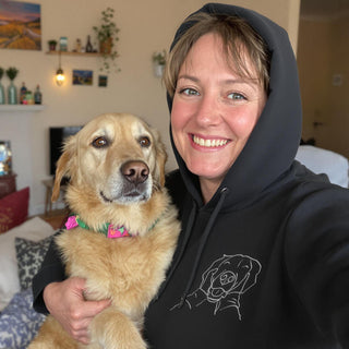 Smiling woman wearing a black hoodie with a line drawing of a dog, holding a Golden Retriever with a pink collar.